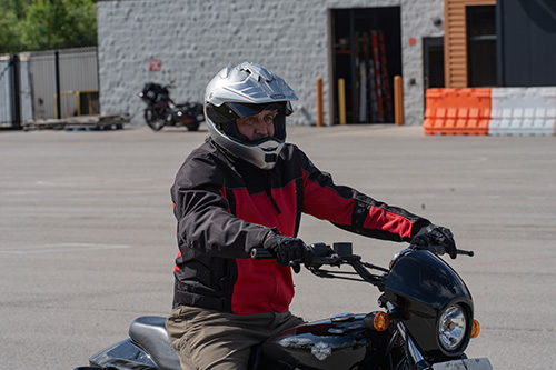 A person sitting atop a motorcycle in a parking lot with orange practice cones in the background.